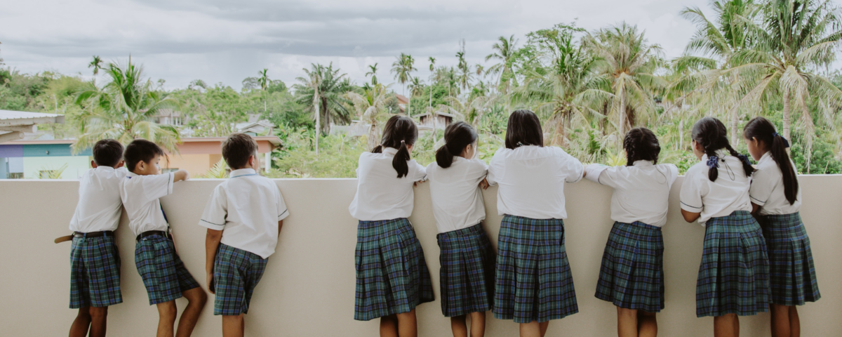 Niños y niñas con vestimenta de escuela, con sus espaldas a la cámara, mirando desde un balcón a un fondo con palmeras